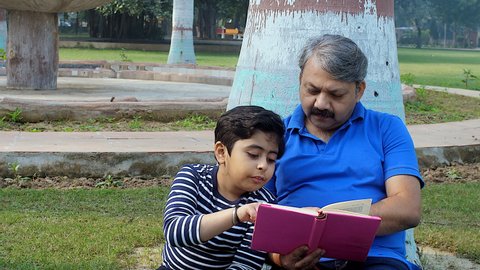 An adorable boy and his grandfather reading a book together in the park. An old man and a little kid sitting together in a public park - leisure time, friendly family, three generations, happy chil...