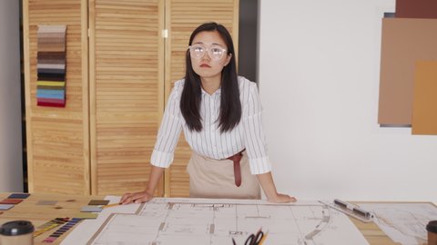 Medium shot of young female Asian architect posing for camera leaning on desk with rolled out house plan and colour and material samples