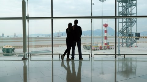Two people stand against glass wall of terminal, woman embrace and lean on boyfriend shoulder. Man look out, silhouetted shot of travelers couple, pensive mood