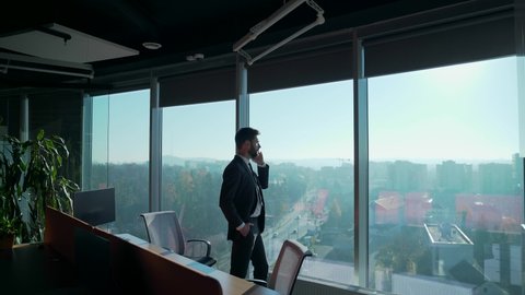 silhouette of a business man in a modern glass office who is talking on a mobile phone on the background of a skyscraper window. Successful confident businessman standing near office window