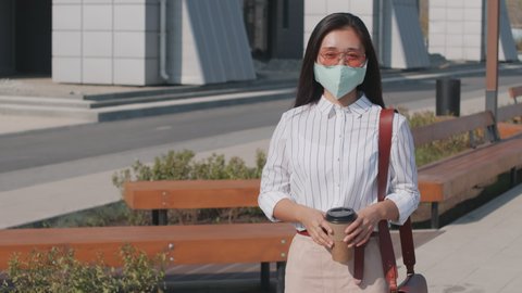 Medium portrait of young Asian woman with takeaway coffee posing for camera outdoors wearing face mask and pink sunglasses