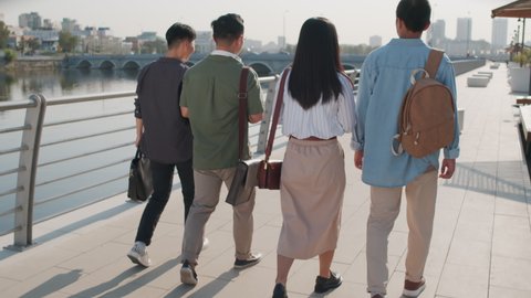 Back-view shot of group of four young Asian people in smart casualwear walking along river embankment on sunny day having conversation