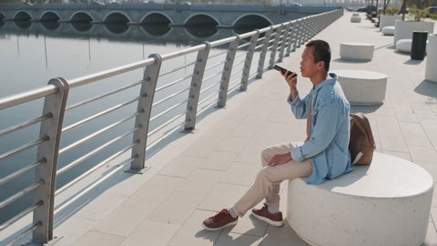 Slowmo shot of young Asian man recording voice message on smartphone sitting at river embankment on sunny day