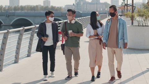 Tracking full-length shot of four Asian friends in face masks talking while strolling along river embankment on sunny day with takeaway coffee