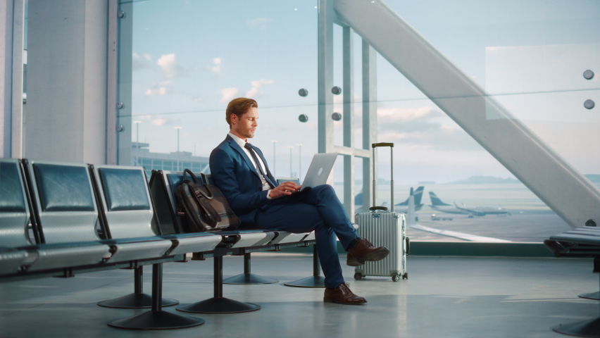 Modern Airport Terminal: Handsome Businessman Working on Laptop Computer While Waiting for His Flight. Man Sitting in a Boarding Lounge of Big Airline Hub with Airplanes Departing and Arriving