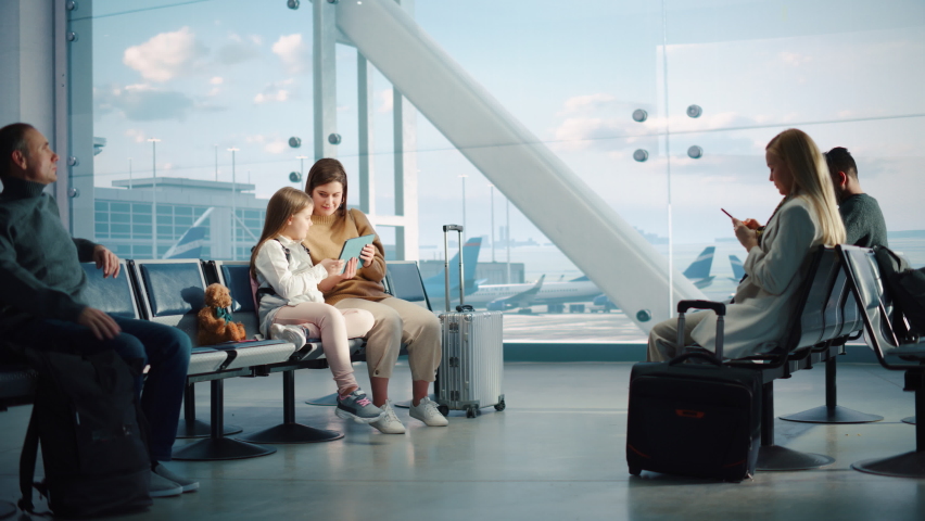 Airport Terminal: Cute Mother and Little Daughter Wait for their Family Vacation Flight, Play Educational Games on Digital Tablet. People Sitting in Boarding Lounge of Airline Hub with Airplanes Fly Royalty-Free Stock Footage #1084638286