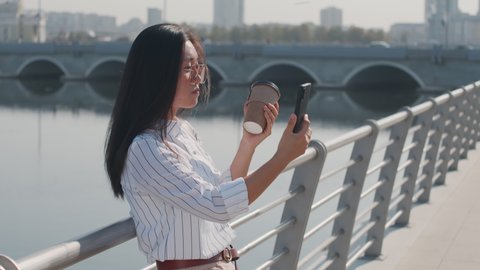 Medium shot of young Asian woman in smart casualwear and sunglasses with takeaway coffee video chatting on smartphone standing at river embankment on sunny day