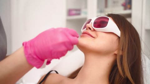 Professional cosmetologist makes a laser facial hair removal to a beautiful woman in a beauty salon. Beautician and spa concept. Body care. Slow motion. Close-up
