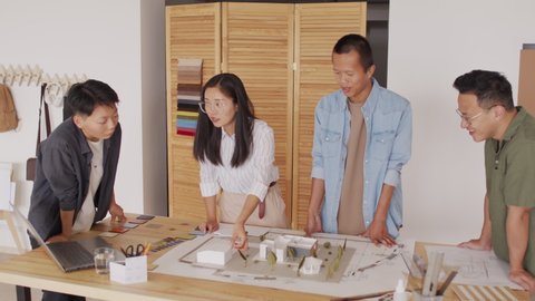 Medium shot of team of four Asian architects looking at house plan and layout on desk, working together at modern office