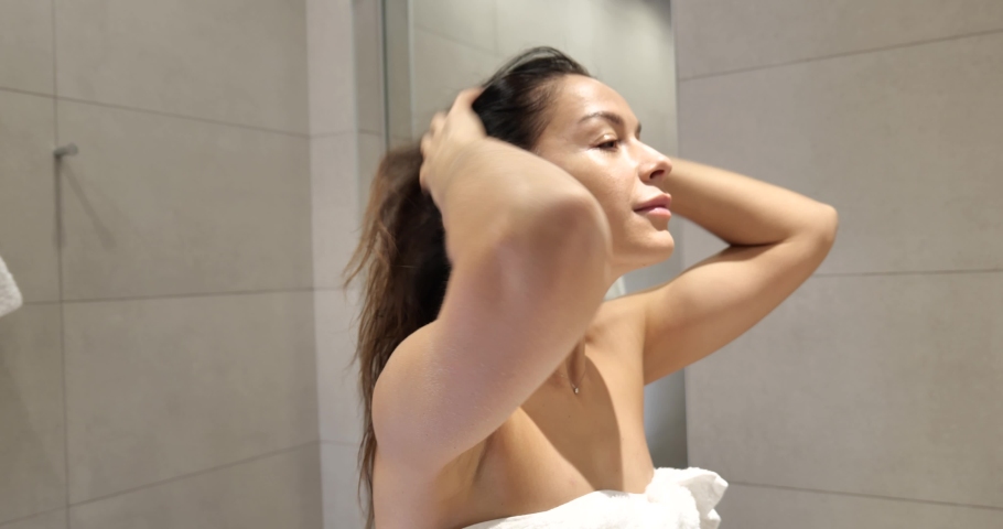 Close up head shot smiling attractive woman looking in mirror, doing morning haircare routine in bathroom. Woman arranging hair, satisfied with strong healthy hair.