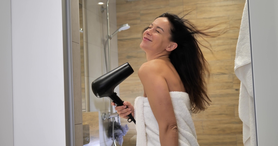 Beautiful woman looking at her reflection in the mirror and smiling while drying hair with blow dryer after taking shower in bathroom. Woman  sings and dances in a shower | Shutterstock HD Video #1084640065