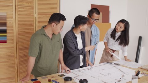 Medium shot of creative young team of four Asian architects working together on housing project, looking at rolled out building plan and colour samples on desk in modern office