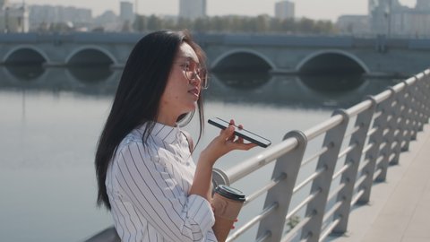 Medium shot of young Asian woman in sunglasses recording voice message on cellphone standing at river embankment on summer day