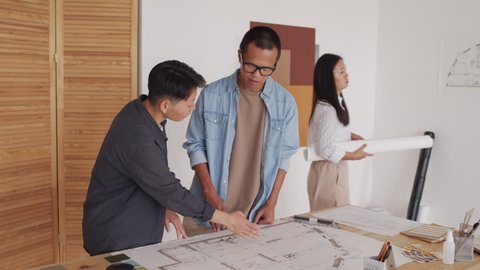 Medium shot of young and creative Asian architects working on housing project together, looking at house plan rolled out on desk at modern office