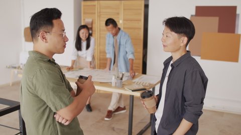 Medium shot of two Asian architects with paper coffee cups in hands having conversation standing at modern office while their colleagues discussing house plan in background