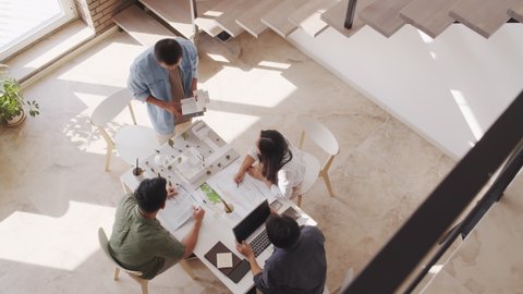 Top-view shot of four Asian architects working in team while discussing housing project looking at its paper layout sitting around table in bright modern office