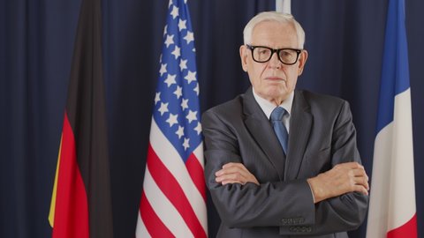 PAN medium portrait of mature male political leader of Caucasian ethnicity posing for camera with hands folded standing on dark blue background with un flags