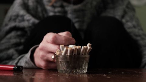Unrecognizable woman smokes, depression concept. Ashtray with cigarette butts and a smoking cigarette in a woman's hand.