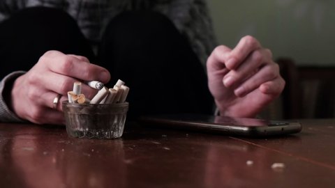 Unrecognizable woman smokes and looks at the phone, depression concept. Ashtray with cigarette butts and a smoking cigarette in a woman's hand.