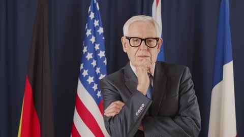 Medium portrait of senior Caucasian male political leader in formalwear and eyeglasses posing for camera standing on dark blue background with un flags
