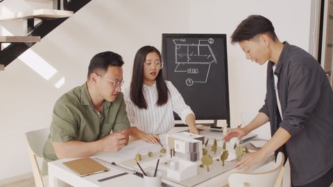 Tracking shot of female Asian architect going down stairs with house layout in hands while working on housing project together with her Asian colleagues
