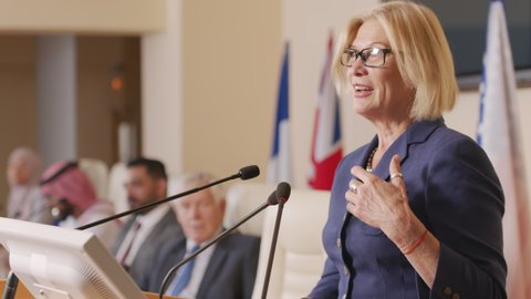 Waist-up shot of mid-adult blonde female politician leader in eyeglasses making speech standing on tribune during official press conference