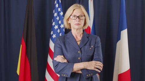 Medium slowmo portrait of mid-adult blonde female politician of Caucasian ethnicity posing for camera in dark blue background with un flags