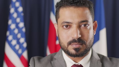 PAN close up portrait of bearded mid-adult politician of Middle-Eastern country looking at camera standing with flags of different countries behind on dark blue background