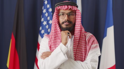 Medium portrait of young male Arab politician in traditional clothing and keffiyeh posing for camera on dark blue background with UN flags