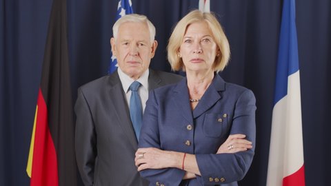 Tilt up medium portrait of couple of Caucasian political leaders posing for camera standing on dark blue background with un flags