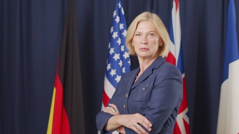 Medium slowmo portrait of Caucasian female president posing for camera standing on dark blue background with UN flags