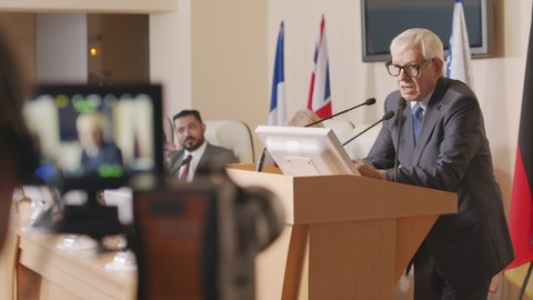 Medium shot of unrecognizable operator recording video of senior Caucasian politician making speech at tribune during press conference