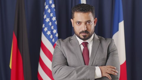 Tilt up medium portrait of serious mid-adult male political leader of Middle-Eastern country posing for camera with hands folded standing on dark blue background with flags of different countries