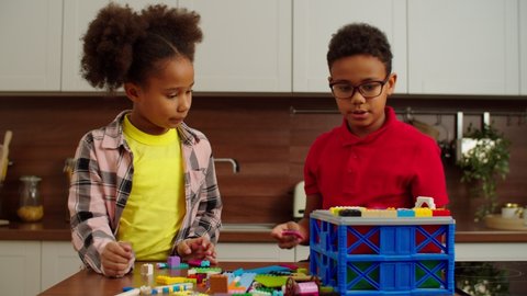 Joyful adorable elementary age black girl and cute preadolescent African American boy in eyeglasses building with colorful kids toy constructor blocks, expressing creativity and imagination at home.