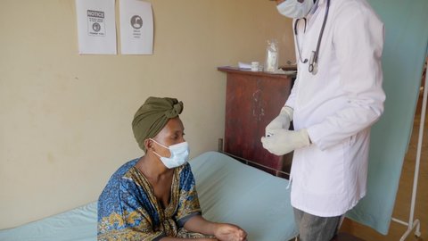 Wide shot of an African male doctor checking a woman with a pulse oximeter in a rural clinic.