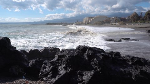 Surf on the sandy beach of the Mediterranean Sea, in the background the Etna volcano, covered with a snow cap in the clouds.  Sicilian coast near Giardini Naxos