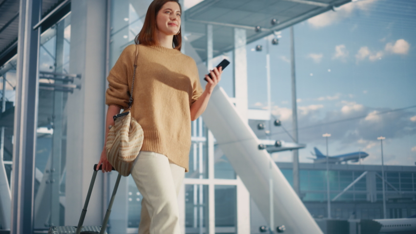 Airport Terminal: Beautiful Smiling Woman Holds Smartphone, Walks in Airline Hub to the Gates Where Airplane Waits Her. Happy Caucasian Female is Ready for Her Flight to Vacation Destination. Royalty-Free Stock Footage #1084670437
