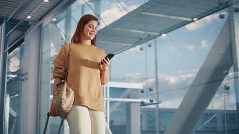 Airport Terminal: Beautiful Smiling Woman Holds Smartphone, Walks in Airline Hub to the Gates Where Airplane Waits Her. Happy Caucasian Female is Ready for Her Flight to Vacation Destination.