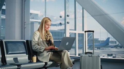 Airport Terminal: Smiling Woman Waits for Flight, Uses Laptop, Browse Internet, does e-Business, Online Shopping. Traveling Female Remote Work Online on Computer in a Boarding Lounge of Airline Hub
