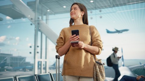 Airport Terminal: Happy Traveling Caucasian Woman Waiting at Flight Gates for Plane Boarding, Uses Mobile Smartphone, Checking Trip Destination on Internet. Smiling White Female in Airline Hub