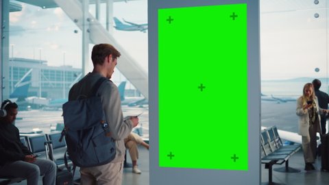 Airport Terminal: Young Man Looking for His Fligt at Green Chroma Key Screen Arrival Departure Information Display. Backgrond: Diverse People Wait for their Flights in Boarding Lounge of Airline Hub