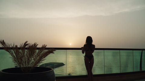 A slender beautiful Caucasian girl in a swimsuit raises her hands up. A European woman stands on the balcony against the backdrop of the sunset and the sea. Lady in bikini on vacation. evening 