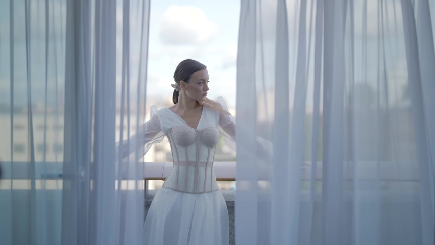 Beautiful woman posing in white dress with corset. Action. Elegant woman inwhite dress poses on terrace. Beautiful view through tulle of posing woman in white dress on terrace