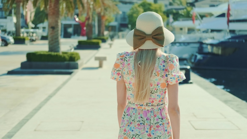 Beautiful blonde woman suddenly turning over her shoulder to the camera and smiling. Walking on the waterfront of the city in summer. Walking in a modern port (marina) among yachts and ships.