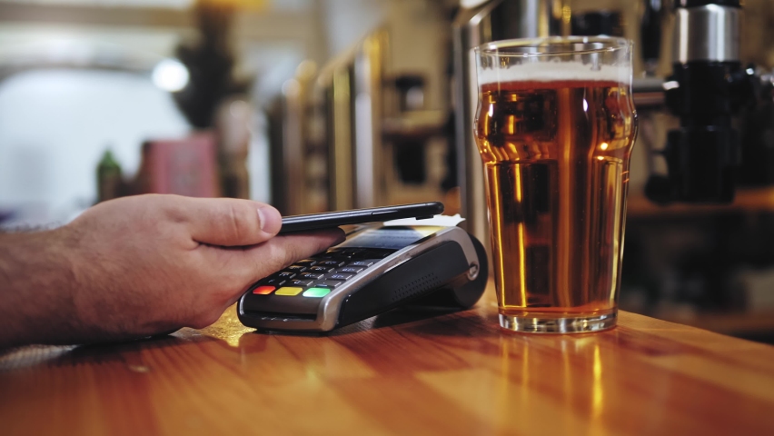 Buying beer and paying by smartphone close-up. Bearded barman standing behind bar counter. Contactless payment in restaurant, touch-free shopping in pubs. | Shutterstock HD Video #1084686097