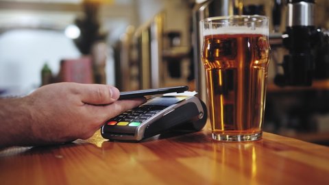 Buying beer and paying by smartphone close-up. Bearded barman standing behind bar counter. Contactless payment in restaurant, touch-free shopping in pubs.