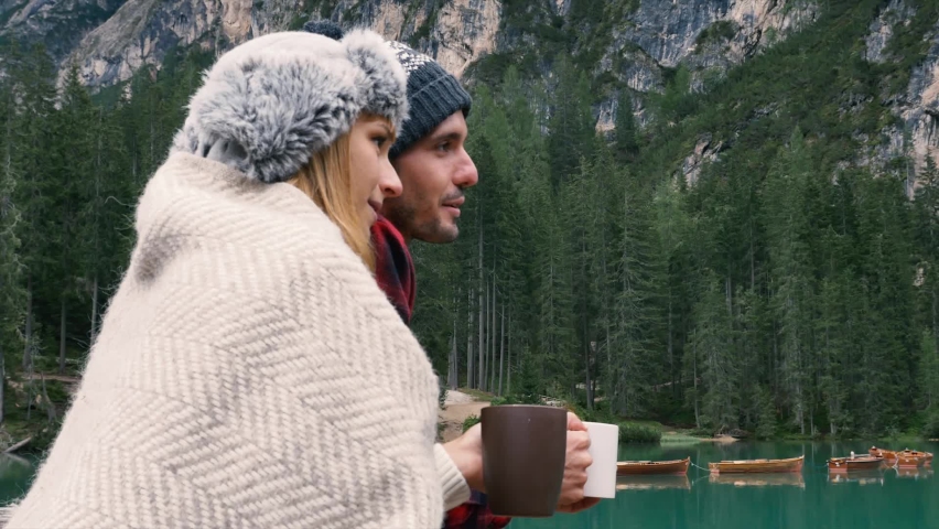 Portrait of a romantic couple of adults visiting an alpine lake at Braies Italy. Tourist in love drinking hot coffee at mountains. Couple, wanderlust and travel concept.