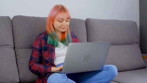 Happy beautiful girl with dyed hair working on laptop computer at home. Young adult woman typing text on notebook on couch. Free lance distant work concept. White female working freelance on lockdown