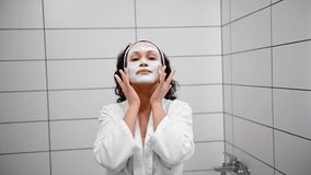 Skin care, beauty home spa concept. Front view of an attractive natural beauty curly-haired woman applying a nourishing alginate mask to her face and making massage movements, cleaning her face.