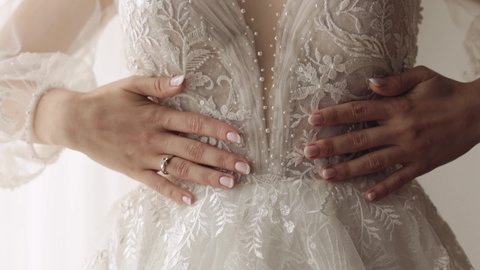 Bride staying near window and touching her white dress. Hands manicure close-up. Engagement ring. Wedding morning preparations. Bride in hotel or home apartment. Woman in gown and veil. Indoors